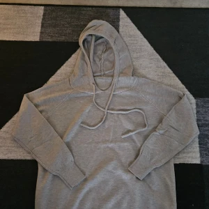 Grå hoodie med snörning - Säljer en enkel och stilren grå hoodie med snörning i huvan. Tröjan har långa ärmar och ribbade muddar vid ärmslut och nederkant. Perfekt för en avslappnad look.
