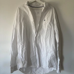 Vit skjorta från 157, storlek XL - Klassisk vit skjorta från 157 i bomull med lång ärm och knappar framtill. Skjortan har en bröstficka med svart broderad detalj och stilren krage med svart kontrastband på insidan. Perfekt för en clean och enkel look.