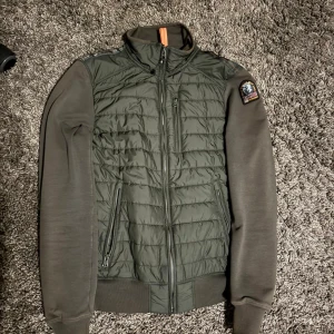 Mörkgrön pufferjacka från Parajumpers - Snygg mörkgrön pufferjacka från Parajumpers med quiltad front och släta ärmar. Jackan har dragkedja, två fickor med zip och coolt patch på axeln. Materialet är polyester och fleece, perfekt för kyliga dagar. Orange detalj i nacken ger extra stilpoäng.