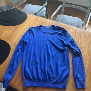 Blå ulltröja från J.Lindeberg - Snygg blå tröja från J.Lindeberg i mjuk ull. Klassisk rund halsringning och ribbade muddar vid ärmslut och nederkant. Perfekt för dig som gillar stilrena och bekväma plagg. Passar till det mesta och är enkel att matcha.