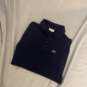 Mörkblå pikétröja från Lacoste - Snygg mörkblå pikétröja från Lacoste med klassisk krage och korta ärmar. Tröjan har knappar framtill och den ikoniska gröna krokodilloggan broderad på bröstet. Tillverkad i mjuk bomull som är skön mot huden. Perfekt för en chill och stilren look.passar 
