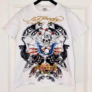 Ed Hardy vit t-shirt - Säljer en vit Ed Hardy t-shirt med stort färgstarkt dödskalle- och panterprint på framsidan, samt guldigt logotryck både fram och bak⭐️Det finns små deoderantfläckar under vardera arm (se bild 4). Axelbredd 37cm, längd 50cm.