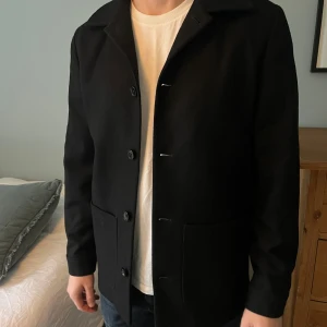 Svart jacka/overshirt med knappar - Stilren svart jacka/overshirt från A Day’s March i ull med klassisk krage och stora framfickor. Lyxig siden på insidan. Jackan har en rak passform och stängs med knappar framtill. Perfekt för dig som gillar minimalistisk och clean look. HELT OANVÄND!! NYPRIS: 2395 kr