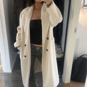Vit lång kappa med knappar - Säljer en stilren och lång vit kappa med bred krage och stora svarta knappar framtill. Jackan har en rak passform och två praktiska fickor på sidorna. Perfekt för dig som gillar minimalistisk och clean stil.