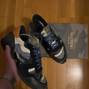 Valentino Rockrunner sneakers camo - Snygga Valentino Rockrunner sneakers med camouflagemönster i svart, grått och beige samt orange detalj vid hälen. Skorna har blå snörning, platt sula och coola nitar på sidan av sulan. Materialmix av skinn och textil ger en unik känsla.