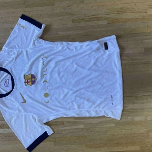 FC Barcelona Lamine Yamal vit matchtröja - Vit FC Barcelona fotbollströja från Nike med mörkblå detaljer vid krage och ärmslut. Tryck med klubbmärke, Nike-logga och månfasmotiv på bröstet. På ryggen står Lamine Yamal och nummer 10 i gult samt UNHCR-logga. Tillverkad i lätt funktionsmaterial.