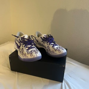 Nike Kobe sneakers vit/lila - Unika Nike Kobe sneakers i vitt med lila detaljer och siffror över hela ovandelen. Skorna har lila snörning, meshmaterial för bra andning och en genomskinlig lila sula med Kobe-logga undertill. Perfekta för dig som vill sticka ut med en sportig och cool look.