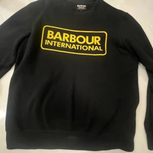 Svart sweatshirt Barbour International L - Svart sweatshirt från Barbour International i storlek L med stor gul logga framtill. Tröjan har rund hals, långa ärmar och är tillverkad i mjuk bomull. Perfekt för dig som gillar streetwear och tydliga loggor.
