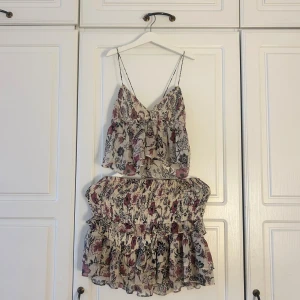 Blommig set med volanger☀️💕💕 - Säljer ett så sött set om är jättepopulärt och finns inte längre att köpa! Blommigt mönster i beige, svart och vinrött. Klänningen har tunna axelband, v-ringad hals och volanger både upptill och nedtill. Materialet är lätt och luftigt, perfekt för varma dagar. Den smockade midjan ger en snygg passform💕💕🌸🥂☀️