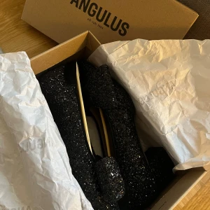 Glittriga svarta pumps från Angulus - Säljer ett par svarta pumps från Angulus med glittrigt material och stor rosett framtill. Skorna har spetsig tå och en stabil, medelhög klack som ger extra attityd. Perfekta för dig som vill sticka ut med en glamorös look.