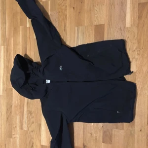 Svart vindjacka från Lacoste med huva - Svart vindjacka från Lacoste med klassisk krokodillogga på bröstet. Jackan har huva, dragkedja framtill och två sidofickor med dragkedja. Perfekt för dig som gillar stilren och sportig look. Materialet är lätt och känns skönt mot kroppen. Nypris 1800