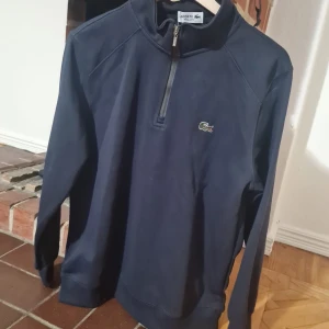 Marinblå half zip tröja från Lacoste - Snygg marinblå långärmad tröja från Lacoste med klassisk krokodillogga på bröstet. Tröjan har half zip-dragkedja och hög krage, perfekt för en stilren och sportig look. Tillverkad i mjuk bomull som känns skön mot huden.