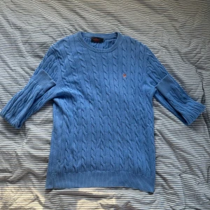 Blå kabelstickad tröja från Morris - Snygg blå kabelstickad tröja från Morris i 100% pima bomull. Tröjan har trekvartsärm, rund halsringning och en broderad orange logga på bröstet. Perfekt för dig som gillar klassisk men ändå chill stil.