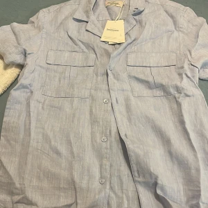 Ljusblå linneskjorta Dressmann S - Fräsch ljusblå skjorta från Dressmann i premium linne. Skjortan har korta ärmar, klassisk krage och två stora bröstfickor med lock. Knappar framtill och en luftig, relaxed passform som är perfekt för varma dagar.