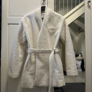 Vit teddyjacka med bälte från NLY Trend - Säljer en fluffig vit teddyjacka från NLY Trend i storlek 34. Jackan har omlottmodell med bälte i midjan, två fickor och en mjuk, mysig textur. 