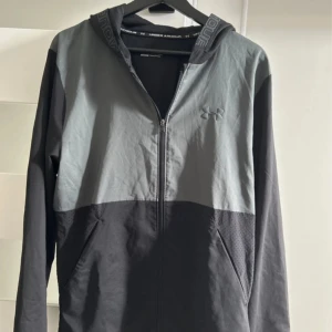 Svart och grå vindjacka Under Armour - Snygg vindjacka från Under Armour i svart och grått med dragkedja framtill och huva med logotypdetaljer. Jackan har långa ärmar, två sidofickor och är tillverkad i lätt polyester som andas.