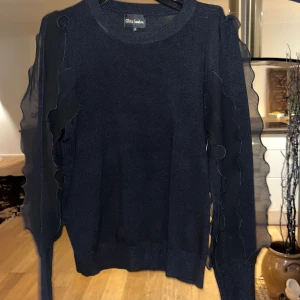 Svart tröja med volangärmar från Rita London - Snygg svart tröja från Rita London med transparenta, vågiga volangdetaljer längs hela ärmarna. Tröjan har rund halsringning och ribbade muddar vid ärmslut och nederkant. Perfekt för dig som vill ha något stilrent men ändå unikt.