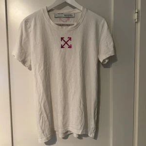Vit Off-White t-shirt med rosa tryck - Vit t-shirt från Off-White med klassiskt rosa pilar-tryck både fram och bak. Rund hals och korta ärmar. Materialet är mjuk bomull och passformen är normal. Perfekt för dig som gillar streetwear och vill sticka ut med en ikonisk design.