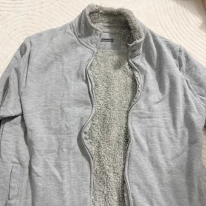 Ljusgrå teddyjacka med dragkedja - Mysig ljusgrå teddyjacka från Nexteria med fluffigt foder och hög krage. Jackan har dragkedja framtill och två sidofickor. Perfekt för kyliga dagar när du vill vara både bekväm och snygg.