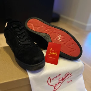 Svarta sneakers från Christian Louboutin - Exklusiva svarta sneakers från Christian Louboutin med klassisk röd sula och snygg snörning. Skorna är tillverkade i mocka och skinn, har rund tå och platt sula. Perfekt för dig som vill sticka ut med en lyxig och stilren look.