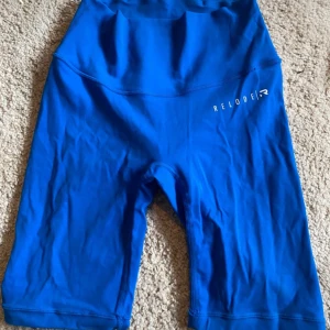 Blå träningstights shorts från Relode - Säljer ett par knälånga, blå träningstights från Relode i storlek XS. De är högmidjade och har en tight passform, perfekta för gymmet eller löpning. Materialet är stretchigt och känns mjukt mot huden. Relode-logga på ena benet. Går också att vika upp om man vill ha kortare, är i mercy kollektionen