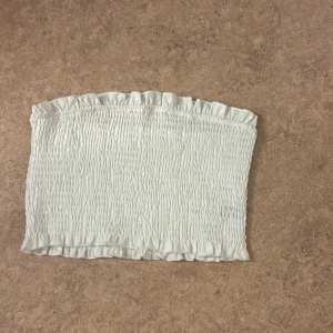 Vit bandeau tubtopp från SHEIN XS - Säljer en vit bandeau tubtopp från SHEIN i storlek Petite XS. Toppen är smockad med volangkant upptill och nedtill, vilket ger en söt och trendig look. Materialet är mjukt och stretchigt, perfekt för varma dagar eller som layering-piece.