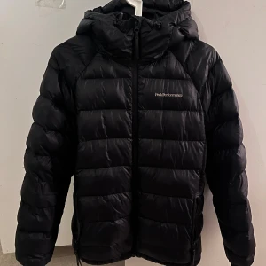 Svart Peak Performance dunjacka - Svart dunjacka från Peak Performance med huva och dragkedja framtill. Jackan är quiltad och har en stilren design med logga på bröstet. Perfekt för kalla dagar och enkel att matcha med olika outfits.