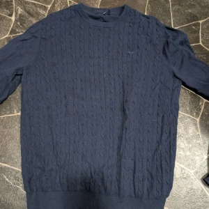 Marinblå stickad tröja Hampton Republ stl Lic - Snygg marinblå stickad tröja från Hampton Republic i storlek L. Tröjan har ett klassiskt kabelstickat mönster och rund hals. Perfekt för dig som gillar stilrena och tidlösa plagg. Mjuk och skön känsla, passar till många olika looks.