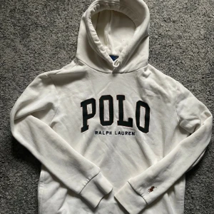 Vit hoodie från Polo Ralph Lauren - Vit hoodie från Polo Ralph Lauren med stor broderad POLO-logga på bröstet och liten brunt broderad häst på ärmen. Tröjan har huva med snörning och är tillverkad i mjuk bomull. Klassisk och clean look med ribbade muddar och lång ärm.