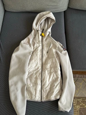 Vit vindjacka från Parajumpers XS - Säljer en snygg vit vindjacka från Parajumpers i storlek XS. Jackan har huva, hel dragkedja framtill, quiltade partier och mesh-liknande ärmar. Detaljer som gul rem vid halsen och logga på ärmen. Perfekt för dig som vill ha en lätt och sportig jacka.