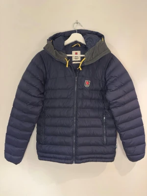 Blå Fjällräven jacka (EXPEDITION PACK DOWN HOODIE) - Säljer en blå Fjällräven Expedition Series dunjacka i storlek S då den inte kommer till användning. Köptes för 4499 på naturkompaniet 2023 och har  använts MAX 10 gånger sedan dess så den är absolut i nyskick. Den enda defekten på jackan är att tvättlappen är avklippt då jag satte en klisterlapp med mitt namn och telefonnummer på som fastnade för hårt och ej gick att ta bort. Pågrund av detta sätter jag ett mycket lågt pris på 2000 kronor för denna jacka. Har drygt 20 bilder till jag kan skicka!