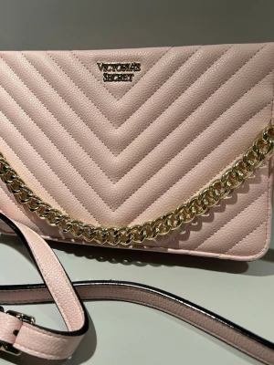Rosa Victoria's Secret Crossbody Väska - Perfekt Skick! - Superfin axelremsväska i ljusrosa med detaljer i guld. Axelremmen är avtagbar och justerbar. Den är quiltad och har en guldfärgad kedja samt en avtagbar läderrem för bekväm användning. Väskan har inga tecken på användning.