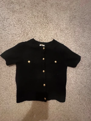 Svart kofta med guldknappar Gina Tricot - Kortärmad svart kofta från Gina Tricot med snygga guldknappar framtill och på fickorna. Koftan är stickad och har en klassisk rund halsringning. Perfekt för lager-på-lager och ger en chic vibe till din outfit.