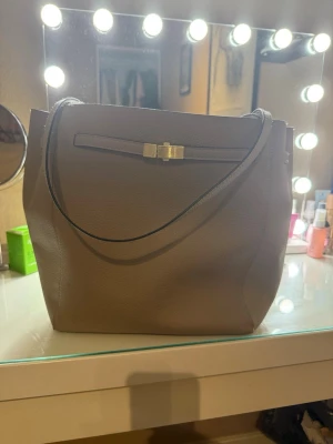 Beige axelväska från H&M i skinnimitation - Snygg beige axelväska från H&M i skinnimitation med stilren design och guldfärgad detalj framtill. Väskan har ett rymligt huvudfack, innerficka med dragkedja och långa handtag som gör den enkel att bära över axeln. Perfekt för dig som vill ha en clean och klassisk look.