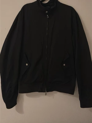 Svart bomberjacka från J.Lindeberg - Säljer en svart bomberjacka från J.Lindeberg i storlek M. Jackan har hög krage, dragkedja framtill och två fickor med tryckknappar. Klassisk och stilren modell med ribbade muddar vid ärmslut och midja. Perfekt för dig som gillar minimalistisk stil. Skick ligger på 7/10, bara att höra av er vid funderingar! 