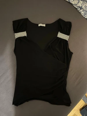 Svart omlottopp med strassdetaljer - Säljer en svart omlottopp från Ayanapa med glittriga strassdetaljer på axlarna. Toppen har en snygg omlott-design och är ärmlös, vilket ger en cool och elegant look. Perfekt för dig som vill sticka ut lite extra.