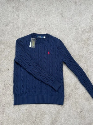 Ralph Lauren Tröja - Säljer en snygg marinblå kabelstickad tröja från Polo Ralph Lauren med klassisk röd logga broderad på bröstet. Oanvänd och alla lappar sitter kvar! Hör av dig vid minsta fråga!