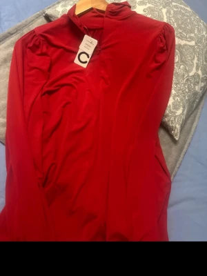 Röd långärmad blus med hög krage - Snygg röd blus med lång ärm och lätt puff vid axlarna. Hög krage och diskret dragkedja framtill. Tunt, mjukt material som faller fint. Perfekt för dig som vill ha en stilren och färgstark topp.