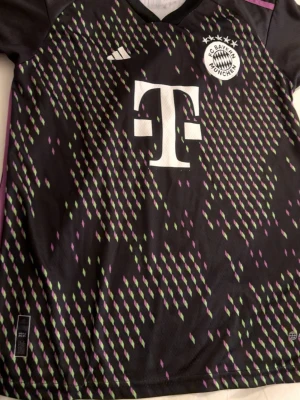 FC Bayern München Kane tröja Adidas - Säljer en FC Bayern München matchtröja från Adidas med Kane och nummer 9 på ryggen. Tröjan är svart med lila och gröna detaljer i ett mönster över hela framsidan, samt stora vita tryck. Perfekt för dig som älskar fotboll och vill ha en unik supportertröja.