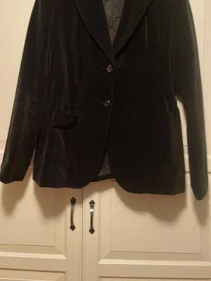 Svart sammetskavaj från Å+ - Snygg svart kavaj i sammetsmaterial från Å+ i storlek 44. Klassisk modell med två knappar och fickor framtill.