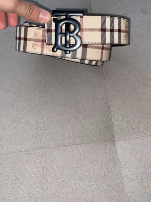 Rutig Burberry bälte med BB-spänne - Säljer ett klassiskt Burberry bälte med det ikoniska rutiga mönstret i beige, brunt och svart. Bältet har ett stort svart BB-spänne framtill och är gjort i skinn. Perfekt accessoar för att lyfta vilken outfit som helst.