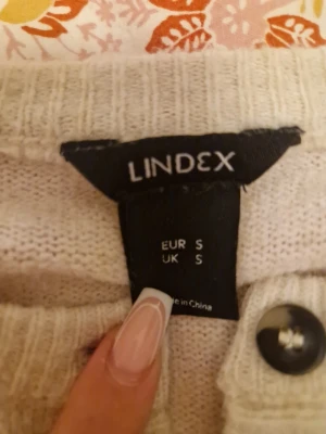 Beige kofta från Lindex med knappar - Mysig beige kofta från Lindex i storlek S. Koftan har långa ärmar, ribbade muddar och knäppning framtill med svarta knappar. Perfekt att slänga på sig när det är lite kyligt ute.