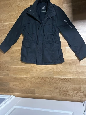 Fieldjacket superdry  - Säljer en svart fieldjacket från Superdry med flera praktiska fickor, hög krage och dragkedja framtill. Jackan har en clean look med diskreta detaljer och patch på ärmen. Perfekt för dig som vill ha en stilren och funktionell jacka till vardags.