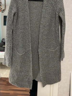 Grå lång kofta från Gina Tricot - Mysig lång stickad kofta från Gina Tricot i grått. Koftan har öppen front, två fickor framtill och långa ärmar. Perfekt att slänga över en t-shirt eller topp under kyliga dagar.