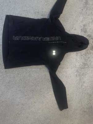 Svart vindjacka från Under Armour - Säljer en svart vindjacka från Under Armour med huva och reflekterande logga på bröstet. Jackan har dragkedja framtill, två sidofickor och tryckt logga längs bröstet. Perfekt för löpning eller utomhusaktiviteter. Materialet är lätt och känns sportigt.