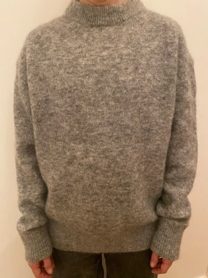 Grå stickad tröja i ullmix - Mysig grå stickad tröja med högre ribbad hals och långa ärmar. Tröjan har en loose passform och breda ribbade muddar vid ärmslut och nederkant. Perfekt för kyliga dagar och enkel att styla till jeans eller kjol.
