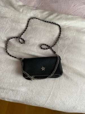 Svart axelväska med kedja och stjärndetalj - Säljer en svart axelväska i skinnimitation från ginatricot,  med silverfärgad kedjerem och en glittrig stjärna framtill. Väskan har dragkedja och en clean, stilren look som passar till allt. Perfekt storlek för det viktigaste och ger en cool vibe till outfiten.