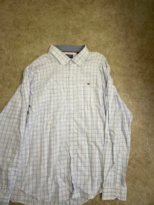 Vit rutig skjorta från Tommy Hilfiger - Klassisk vit skjorta från Tommy Hilfiger med ljusblå rutmönster och diskret logga på bröstet. Skjortan har lång ärm, knappar hela vägen och en snygg krage. Perfekt till jeans eller chinos för en clean look.