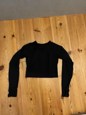 Svart croppad långärmad topp - Säljer en svart croppad topp med långa ärmar och rund hals. Toppen har ribbade detaljer på sidorna och baktill som ger en sportig känsla. Perfekt att styla med höga jeans eller kjol för en clean look.