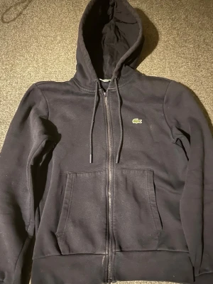 Lacoste zip hoodie - En mörkblå Lacoste zip hoodie som inte använts speciellt mycket och är i ett bra skick. Den är i storlek XS och jag säljer den för jag har vuxit ur den.